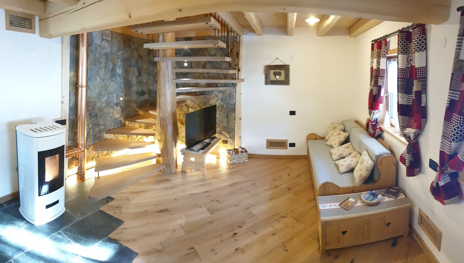 Soggiorno con stufa bianca, scala a chiocciola in legno su parete di pietra e divano con cuscini a cuore